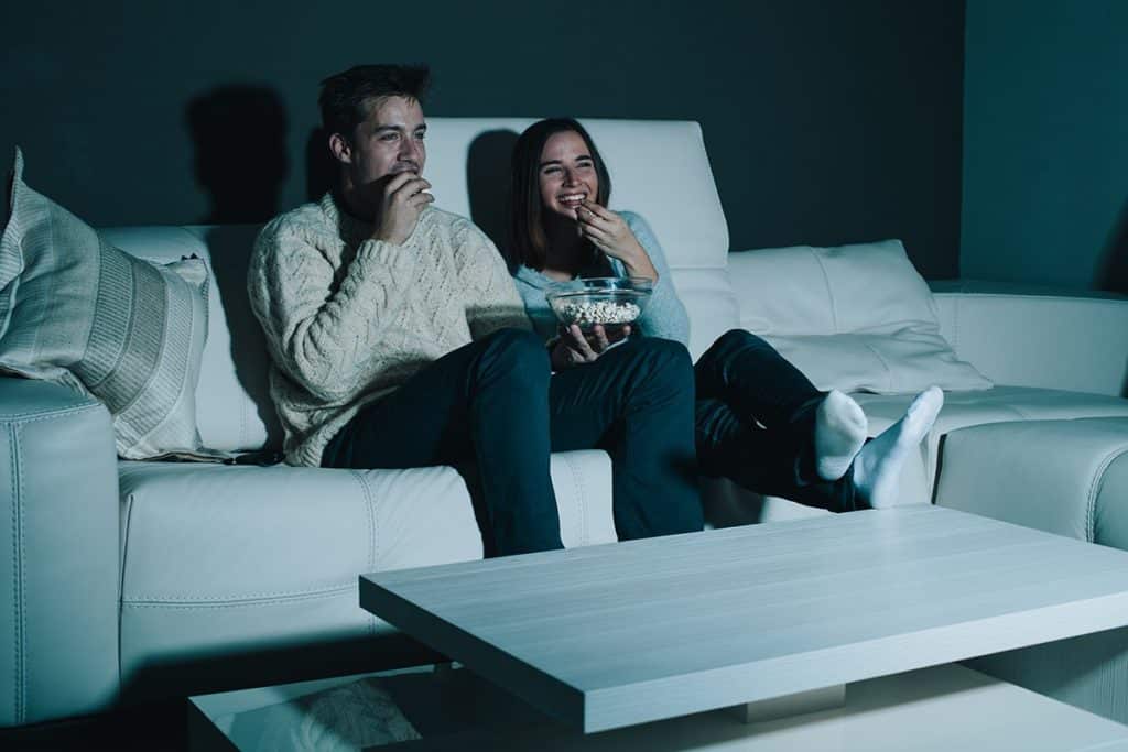 A man and woman watch television on a couch while eating popcorn