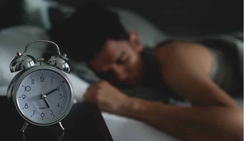A man rests with an alarm clock on the nightstand.