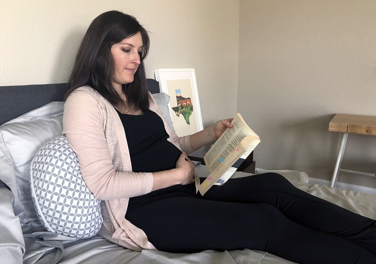 a woman uses the boppy side sleeper pillow as an arm rest, while she reads a book in bed