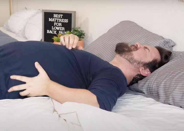 A man laying on a bed clutches his lower back and shows he is in pain