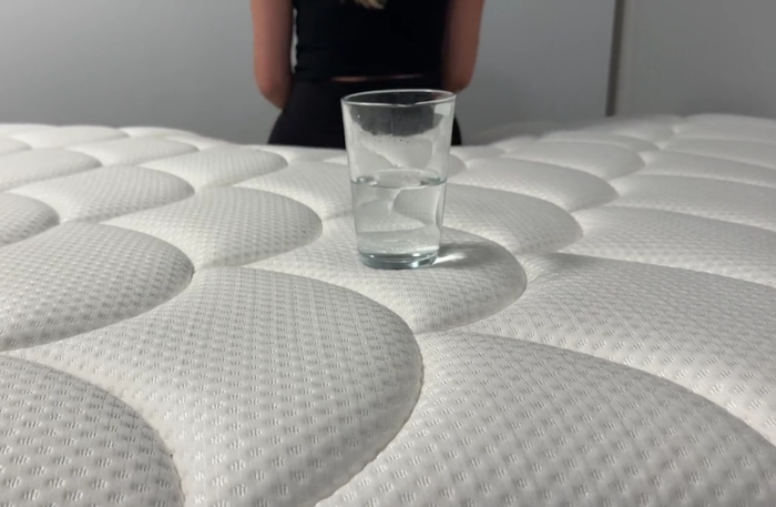 A person sits on a DreamCloud Classic Hybrid while a glass of water is monitored on the mattress.