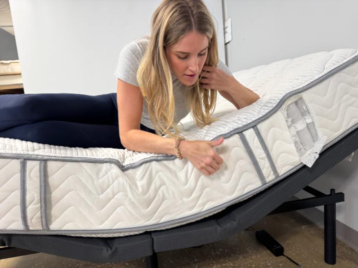 Kayla examining the side of the DLX Classic Two-Sided Mattress while it's on an adjustable base, with a cutaway view of its springs near the head of the bed