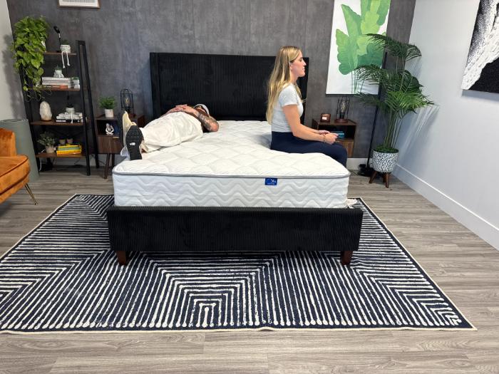 Kayla sitting up on one side of the DLX Classic Two-Sided Mattress while Sosha lies blindfolded on the other side